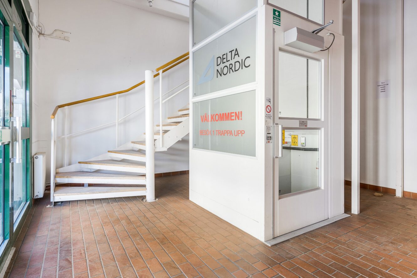En liten hiss och en vit spiraltrappa i en byggnadslobby med tegelgolv och glasdörrar. Skyltar på hissen visar "Delta Nordic" och svensk text.