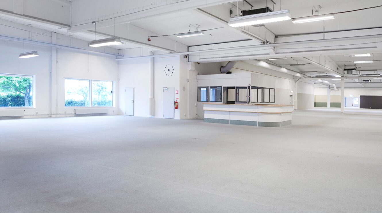 Rymlig, tom interiör i en kommersiell byggnad med vita väggar, stora fönster, synliga rör i taket och en central receptionsdisk.