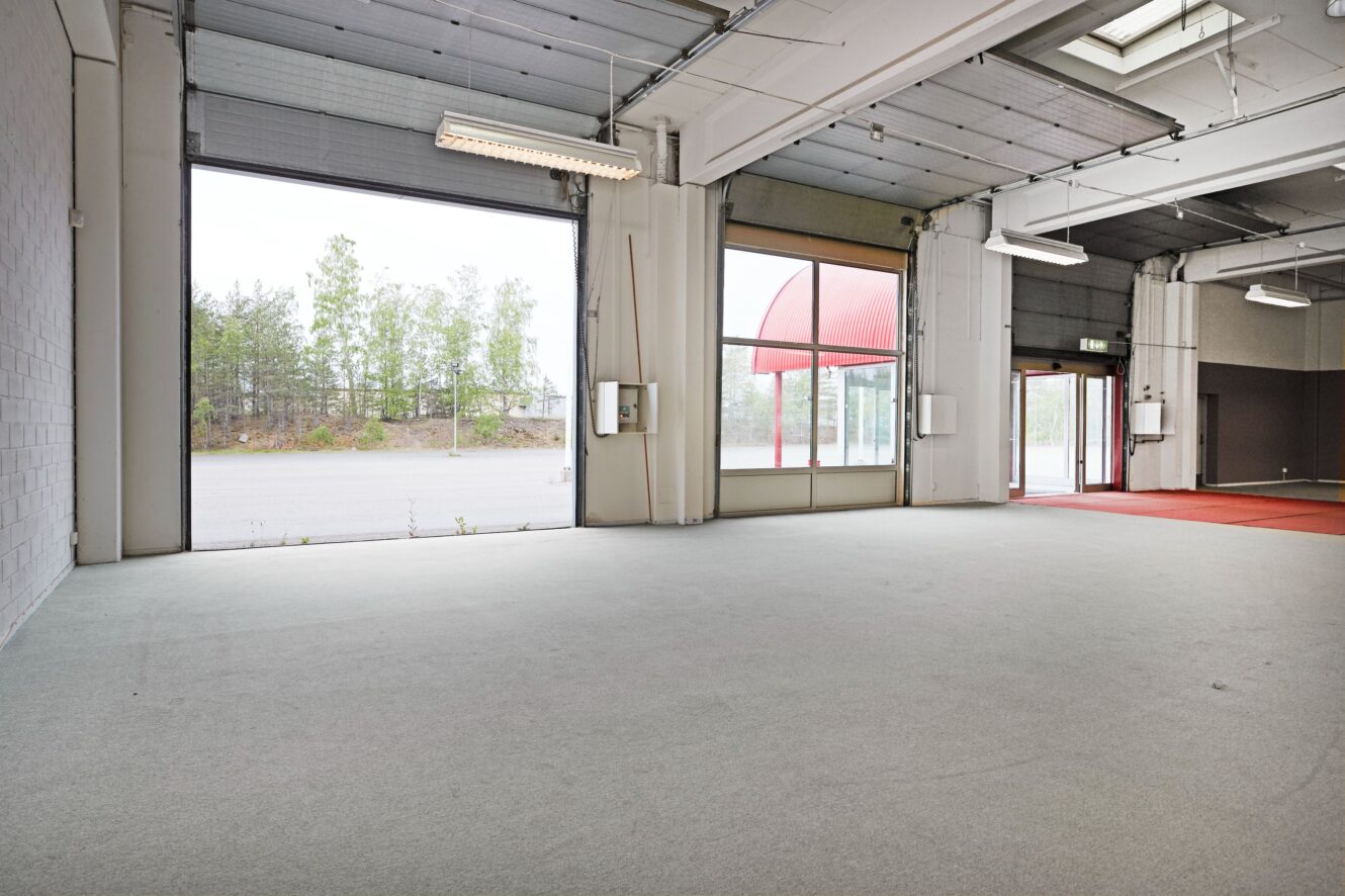 Tom affärslokal med stora fönster, grå heltäckningsmatta, högt i tak och synlig parkeringsplats utanför; väggarna är neutrala, med takbelysning och synliga rör.