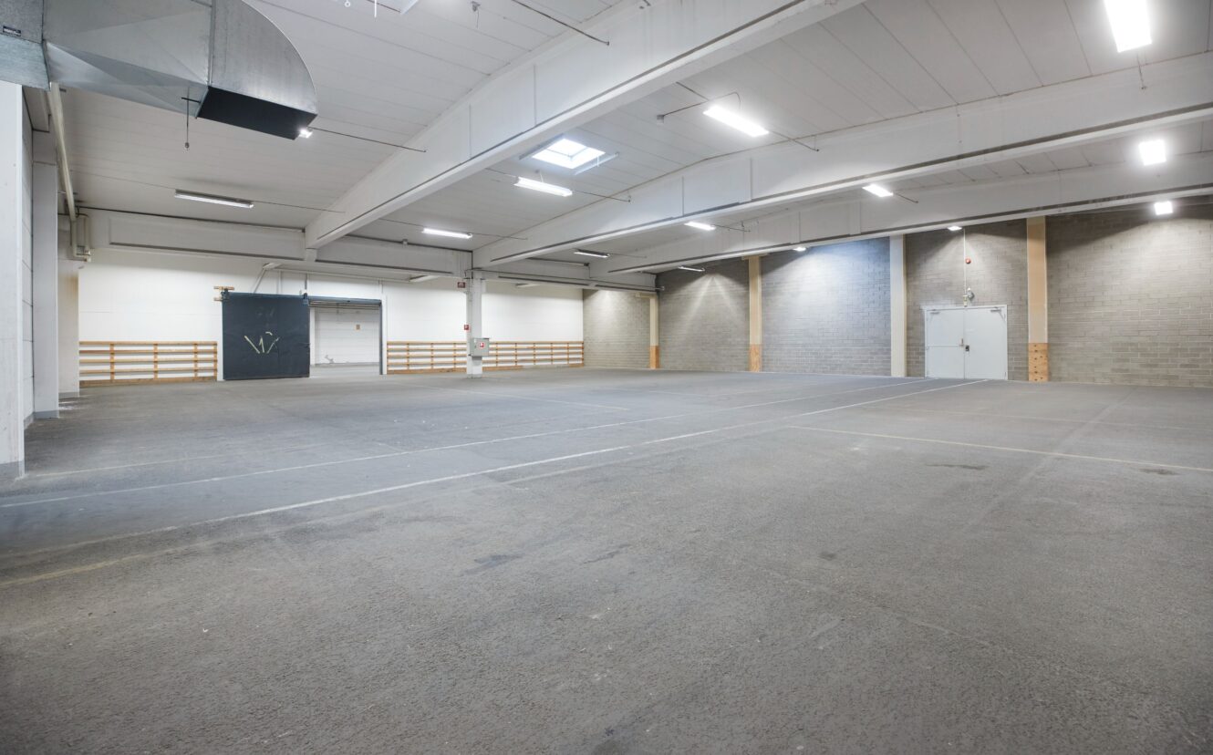 Ett stort, tomt industrilager med betonggolv, högt i tak, takbelysning och några dörrar längs bak- och sidoväggarna.