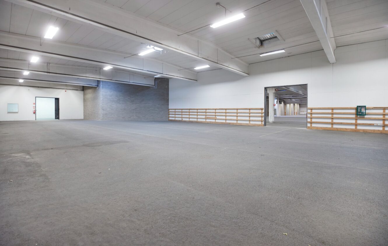 Invändig vy av ett stort, tomt industrilager med vita väggar, takbelysning och betonggolv. Flera dörröppningar och ett räcke kantar den högra sidan.