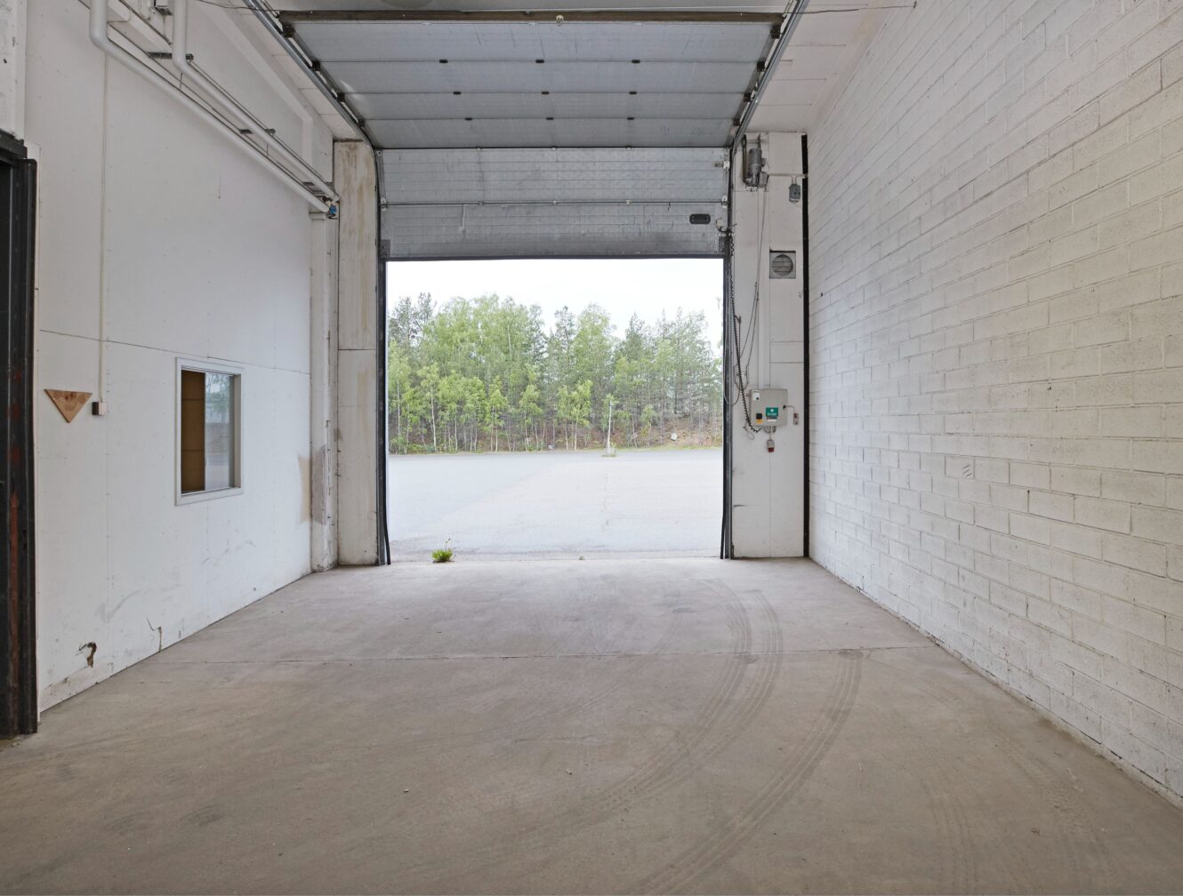 Ett tomt industrigarage med en upphöjd rullport, vita tegelväggar, betonggolv och en yttre vy med träd och trottoar.