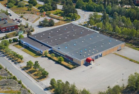 Flygfoto över en stor industrihall med en röd markis över entrén, omgiven av en tom parkeringsplats och träd.