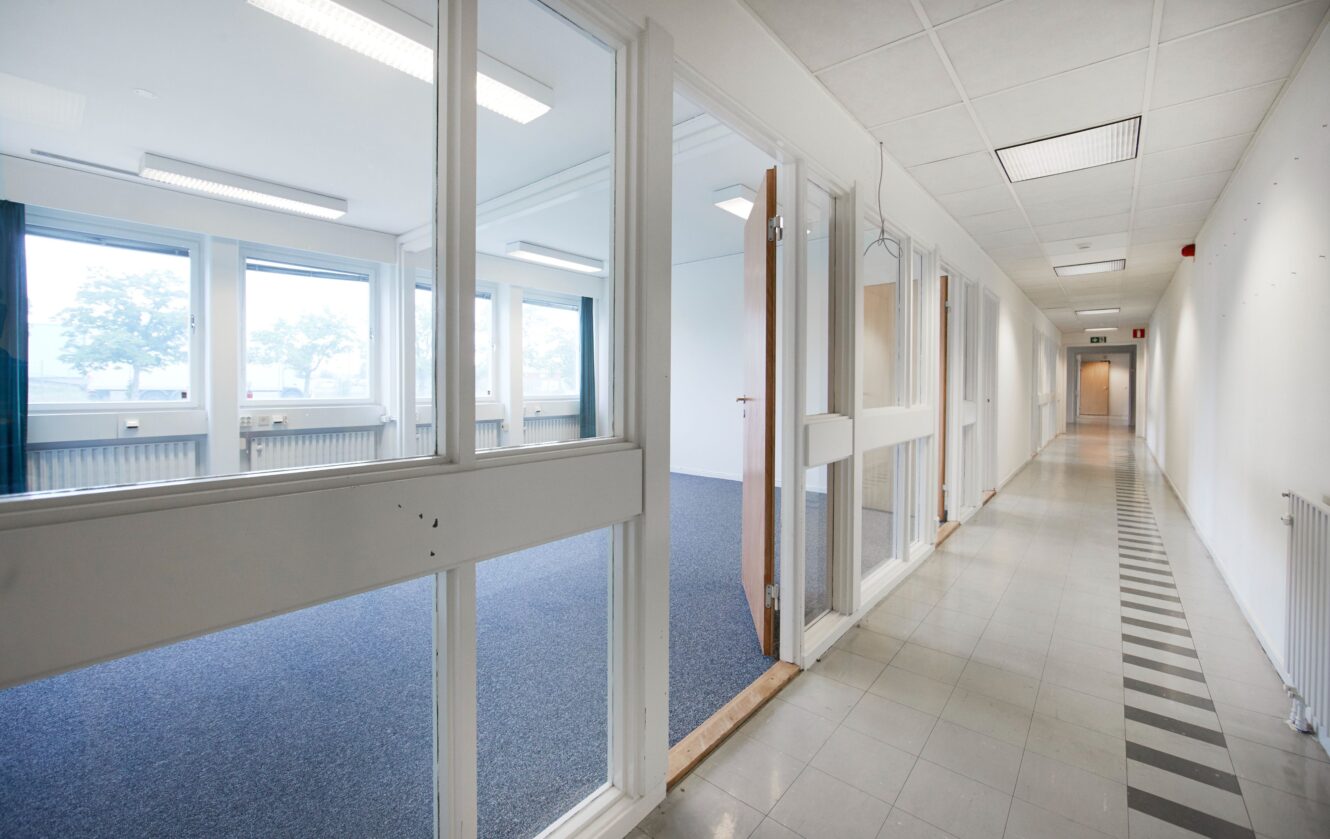 En lång, ljus korridor med vita väggar och klinkergolv, intill ett tomt kontorsrum med stora fönster och glaspartier.