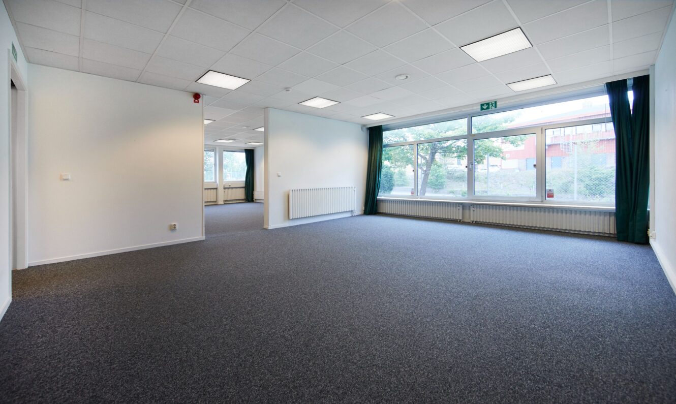 Rymligt, tomt kontorsrum med grå matta, vita väggar, stora fönster och taklampor.