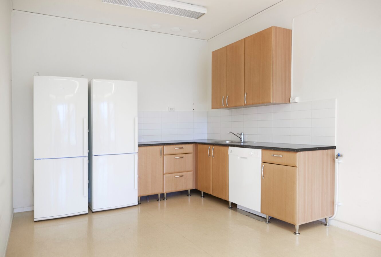 Ett enkelt kök med två vita kylskåp, träskåp, en diskho, en diskmaskin, kaklat stänkskydd och beige golv.