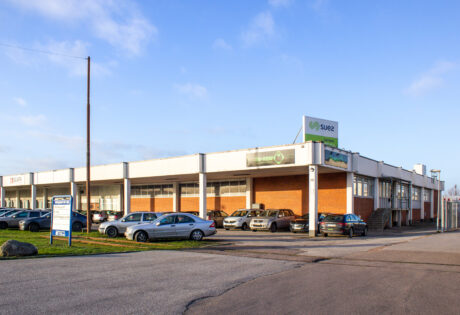 Envånings affärsbyggnad med stora fönster, fasad i tegel och vitt och flera parkerade bilar framför. En SUEZ-skylt är synlig på taket.