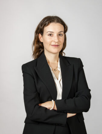 En kvinna i svart kavaj och vit blus står med armarna i kors och bär ett halsband och ett armband mot en ljus bakgrund.