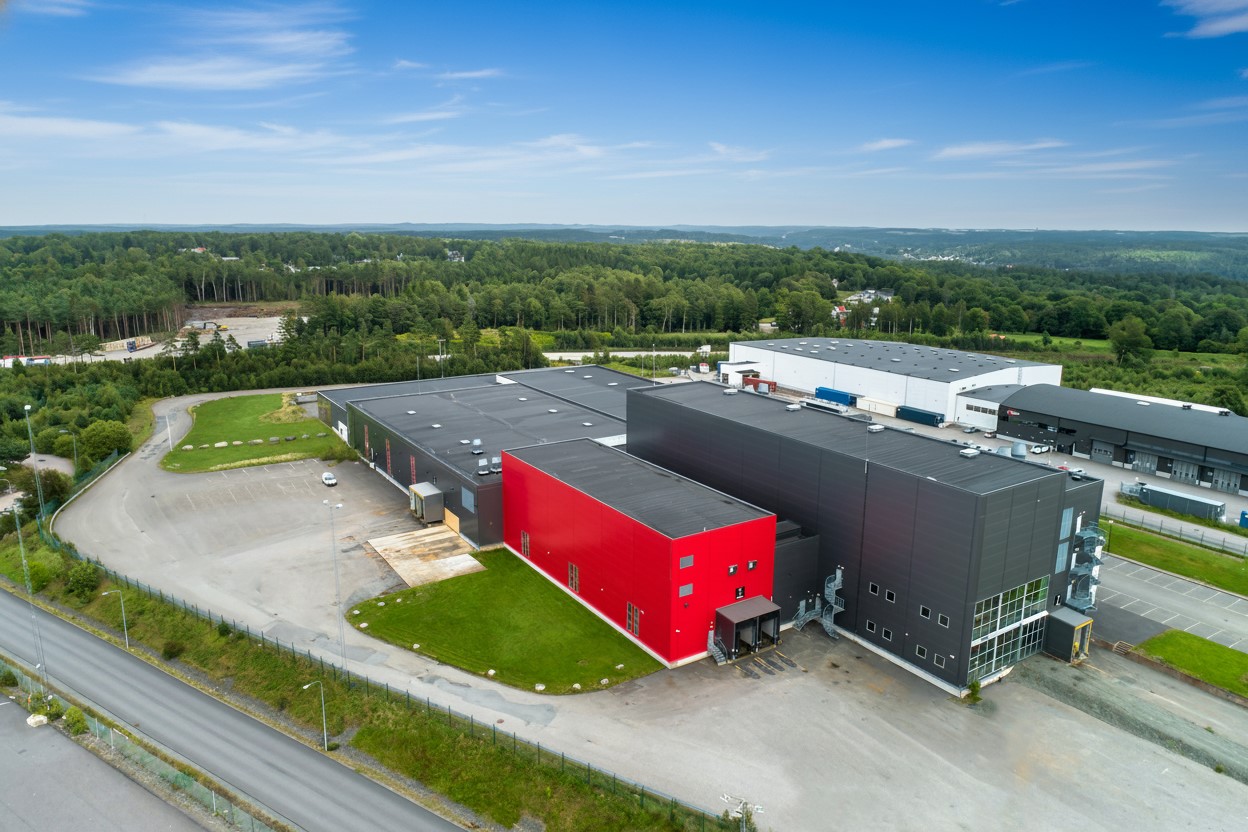 Flygfoto över ett modernt industriellt lagerkomplex med svart och röd exteriör, omgivet av parkeringsplatser och grönska.