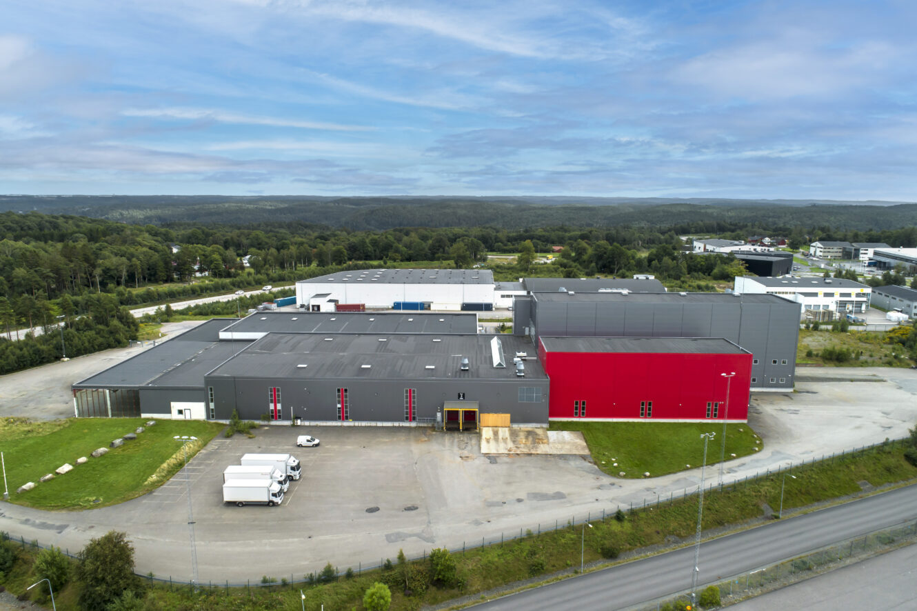 Ett stort industriellt lager med en röd och svart mönstrad sektion, omgiven av träd och andra byggnader, med lastbilar parkerade utanför.