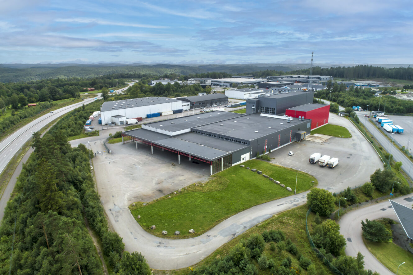 Flygfoto över ett stort industriellt lager med en röd sektion, omgiven av grönska, parkeringsplatser och närliggande vägar. Flera lastbilar står parkerade vid lastkajer.