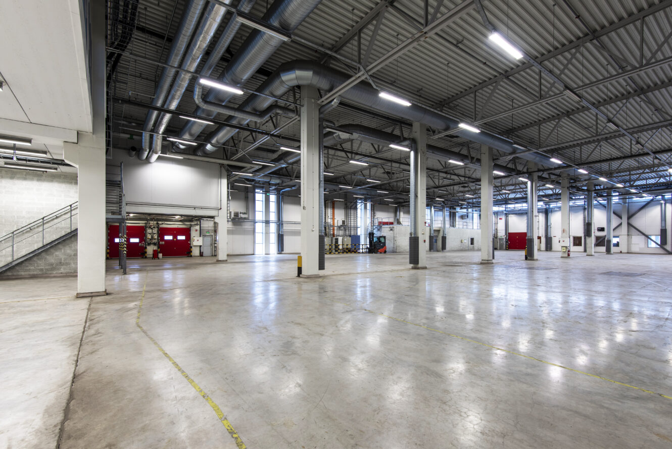 Stort, tomt industrilager med polerade betonggolv, synliga rör i taket, stödpelare och lastkajer längst bort. Utrymmet är upplyst av lysrör.