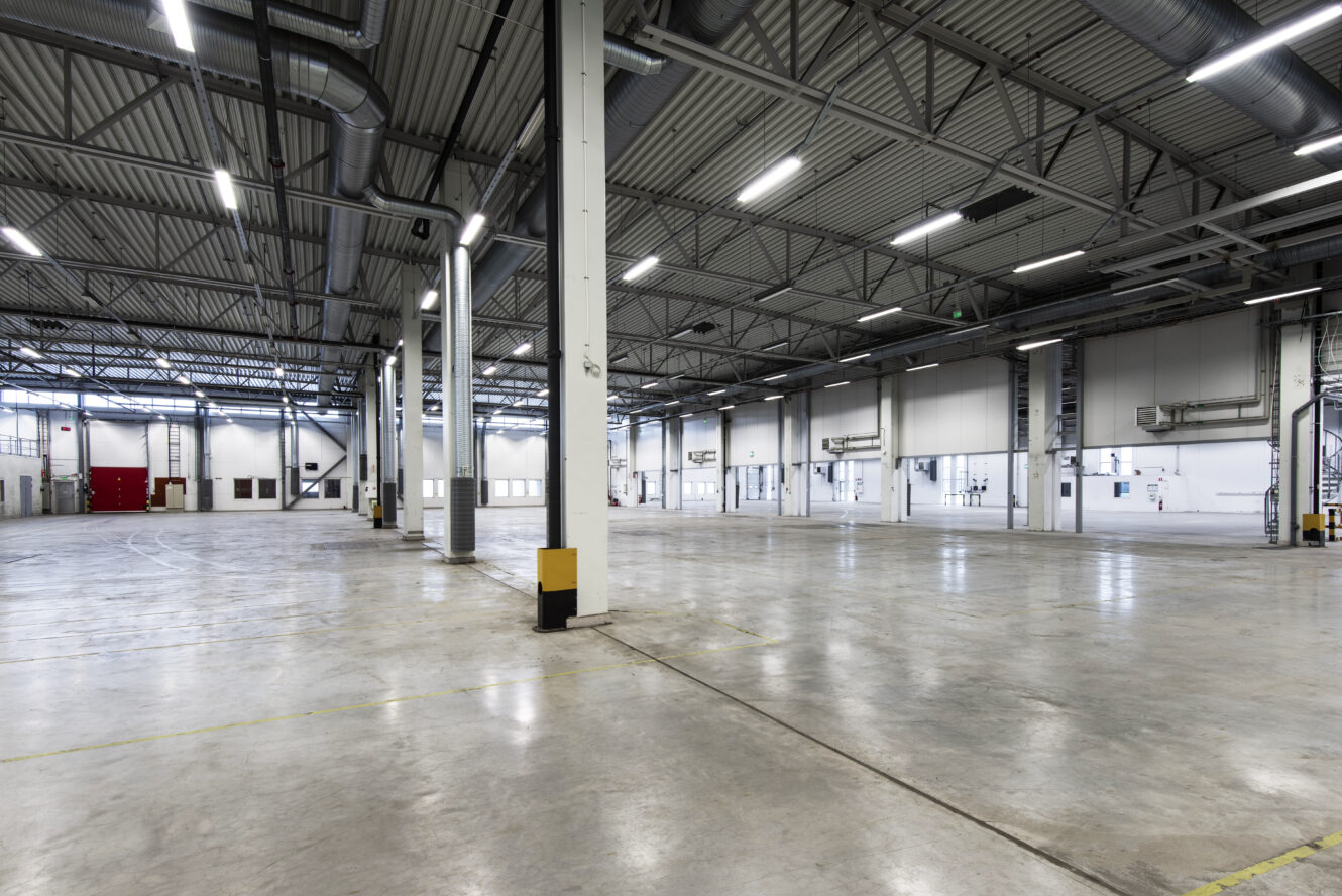 Interiör i en stor, tom industrilokal med högt i tak, synliga metallbalkar, takbelysning och betonggolv.