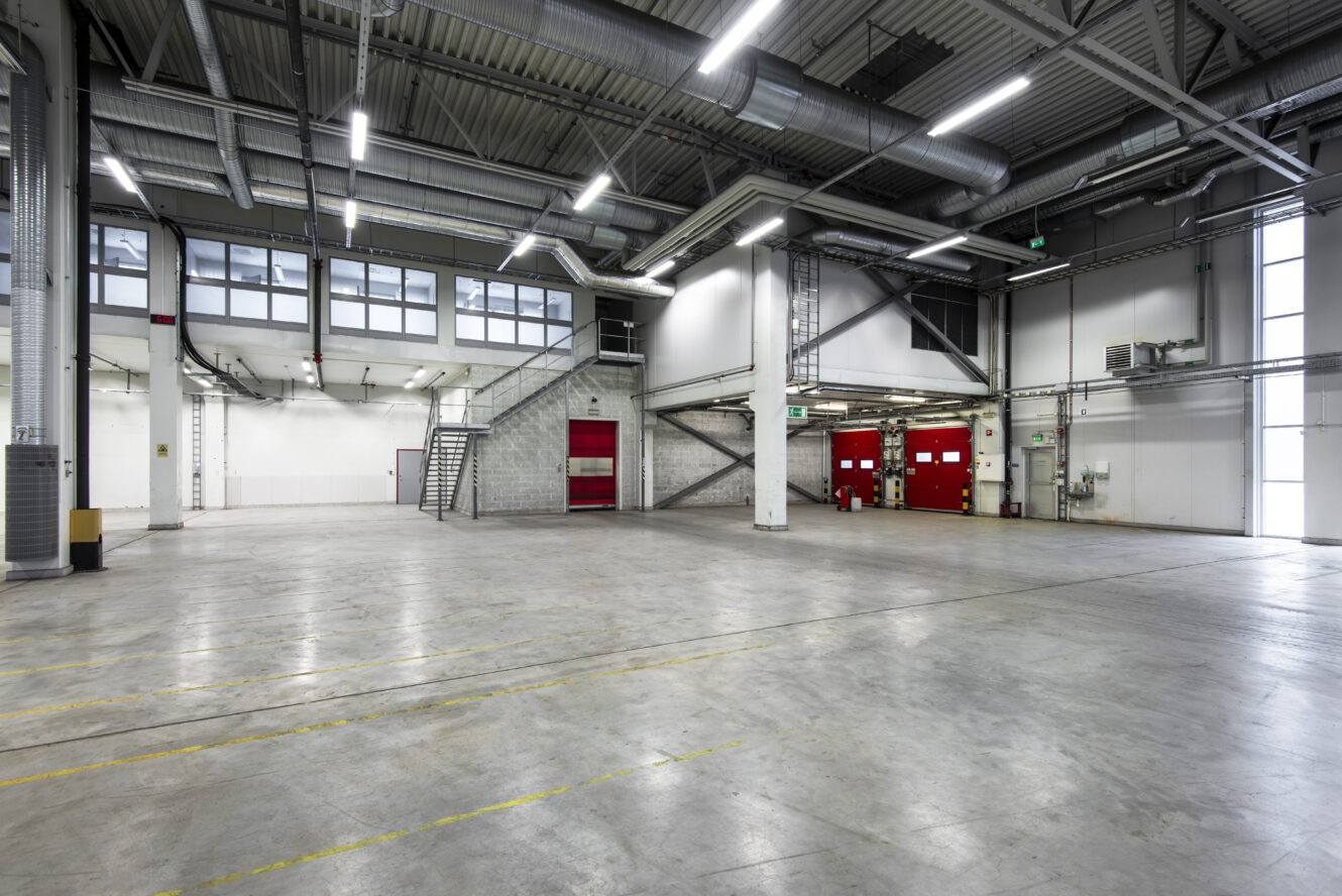 Rymlig, tom lagerlokal med betonggolv, högt i tak, industriell belysning, stora fönster, röda lastbryggor och metalltrappor.