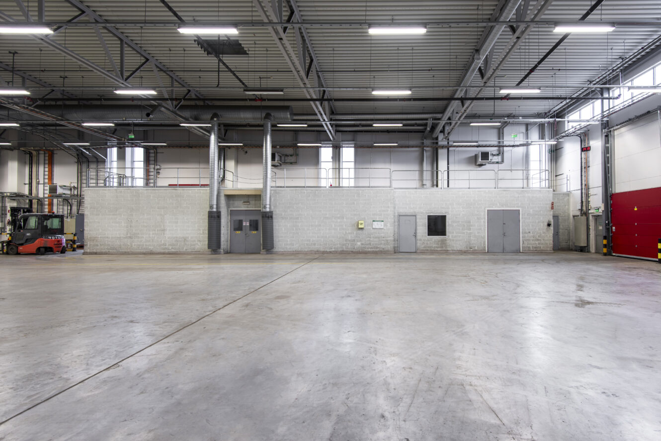 Stor tom lagerlokal med betonggolv, högt i tak, industribelysning, ett litet slutet kontorsutrymme och en gaffeltruck parkerad på vänster sida.