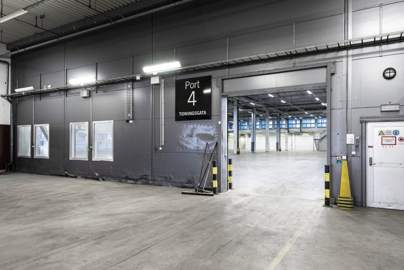 Interiör i en tom industrihall med högt i tak, stora fönster och en skylt med texten Port 4 ovanför en bred ingång till ett öppet utrymme.