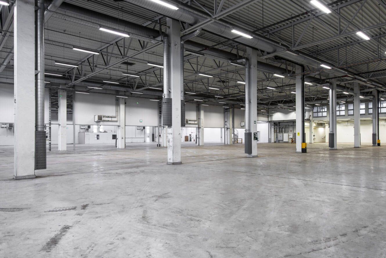 Stor, tom industrilagerlokal med betonggolv, högt i tak, stödpelare och takbelysning.