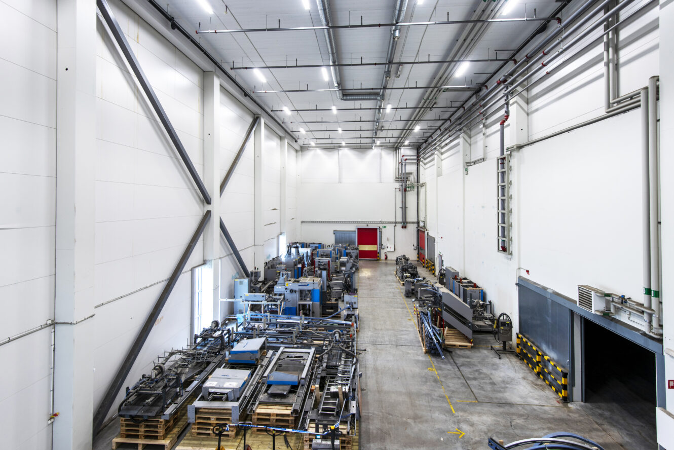 Stor industriell lagerlokal med maskiner och utrustning placerade på pallar längs båda sidor av en bred mittgång under stark takbelysning.