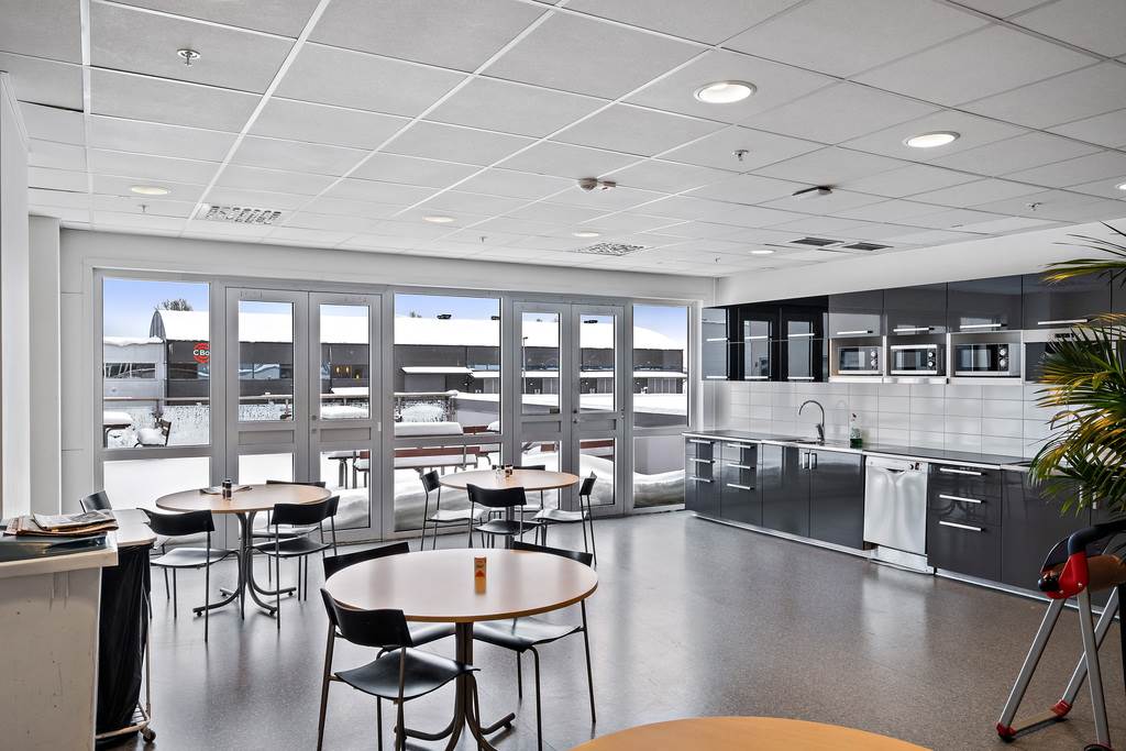 Modernt fikarum med runda bord, svarta stolar och ett pentry med mörka skåp och inbyggda vitvaror. Stora fönster visar en snöig utomhusscen med byggnader.