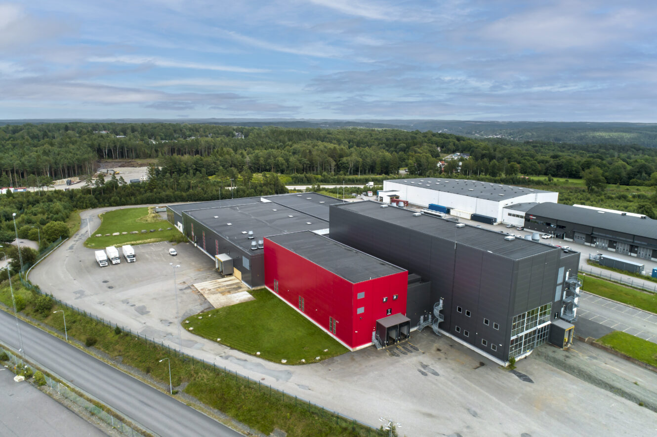 Flygfoto över en modern industribyggnad med en distinkt röd fasad, omgiven av parkeringsplatser och grönska, belägen nära ett skogsområde.