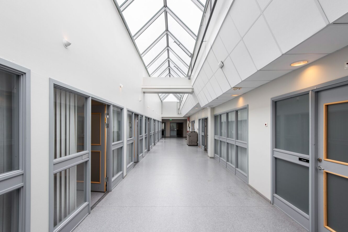 En väl upplyst hall med glasdörrar på båda sidor, ett ljusgrått golv och ett snedställt glastak som släpper in naturligt dagsljus.