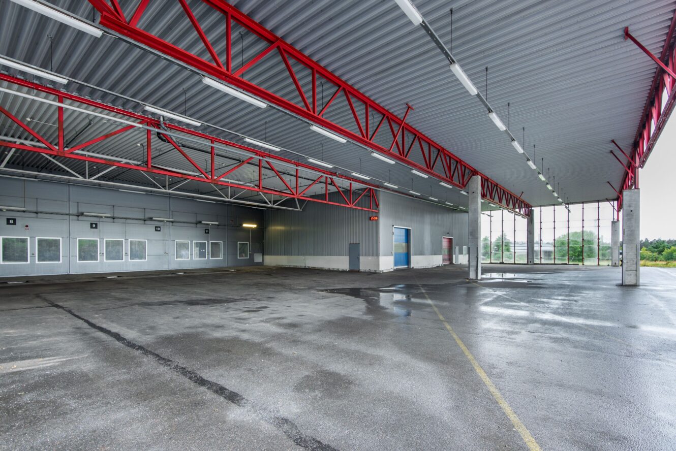 Stort tomt industrilager med vått betonggolv, röda takbjälkar, flera fönster och öppna väggar som avslöjar grönska utanför.