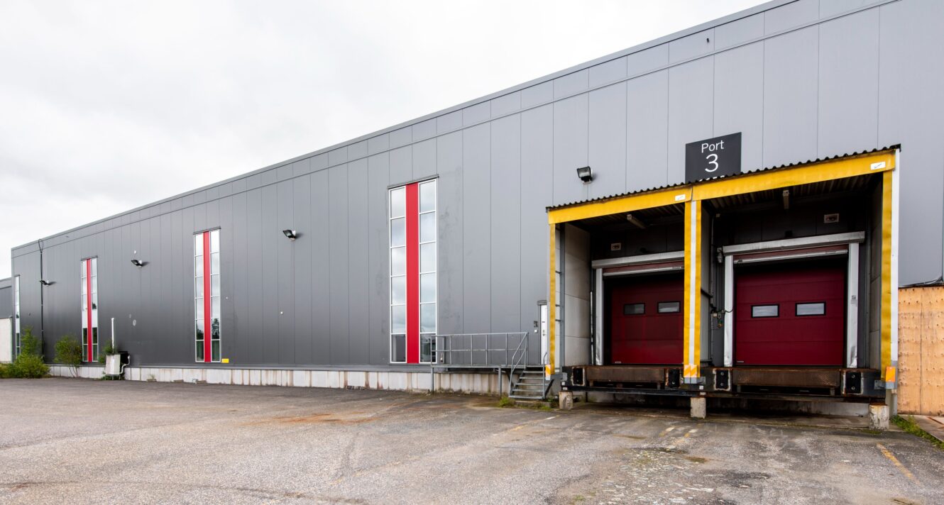 Grå industrilagerfasad med två röda lastkajdörrar märkta Port 3, höga fönster och en asfalterad yta i förgrunden.