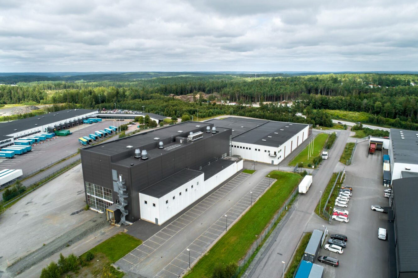 Flygfoto över ett stort industriellt lagerkomplex med parkeringsplatser, leveransbilar och omgivande grönska under en molnig himmel.