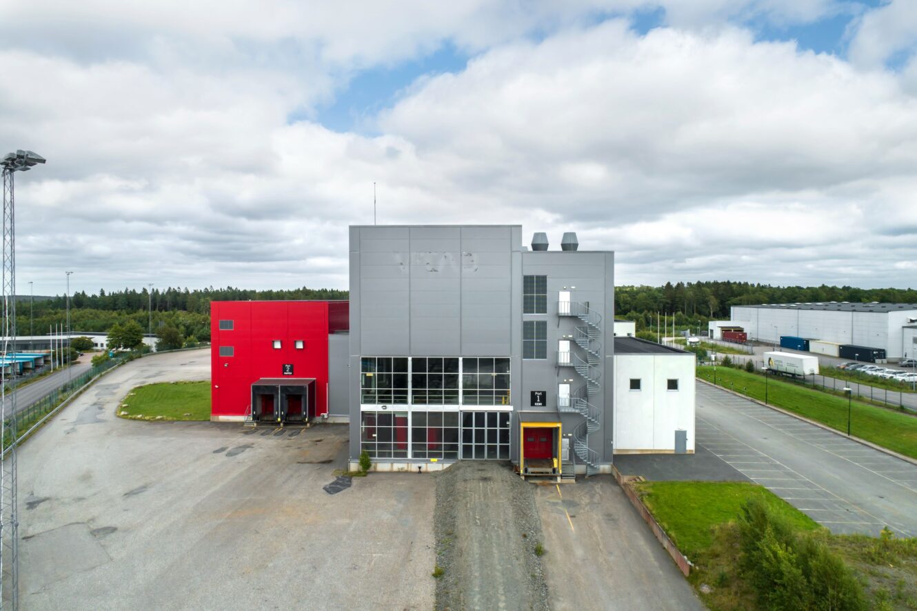 En modern industribyggnad med röda och grå sektioner, lastkajer och utvändiga trappor, omgiven av parkeringsplatser och grönska under en molnig himmel.