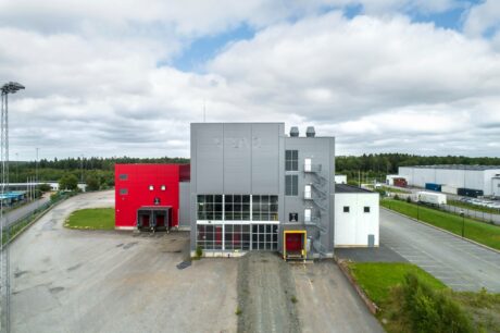 En modern industribyggnad med röda och grå sektioner, lastkajer och utvändiga trappor, omgiven av parkeringsplatser och grönska under en molnig himmel.