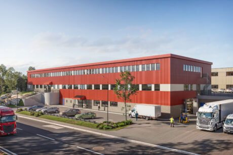 En modern industribyggnad i två våningar med röd fasad, lastbilar parkerade utanför och människor som arbetar vid lastområdet intill en huvudväg.
