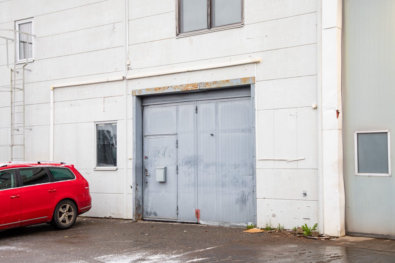Stor grå industriport i metall på en vit husvägg, bredvid en röd bil som står parkerad på en våt trottoar.
