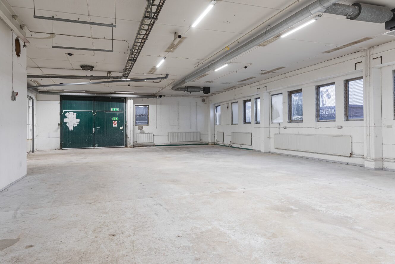 Tomt industrirum med vita väggar, rör i taket, lysrör och en grön dubbeldörr längst bort. Flera fönster längs den högra väggen. Golvet är bar betong.