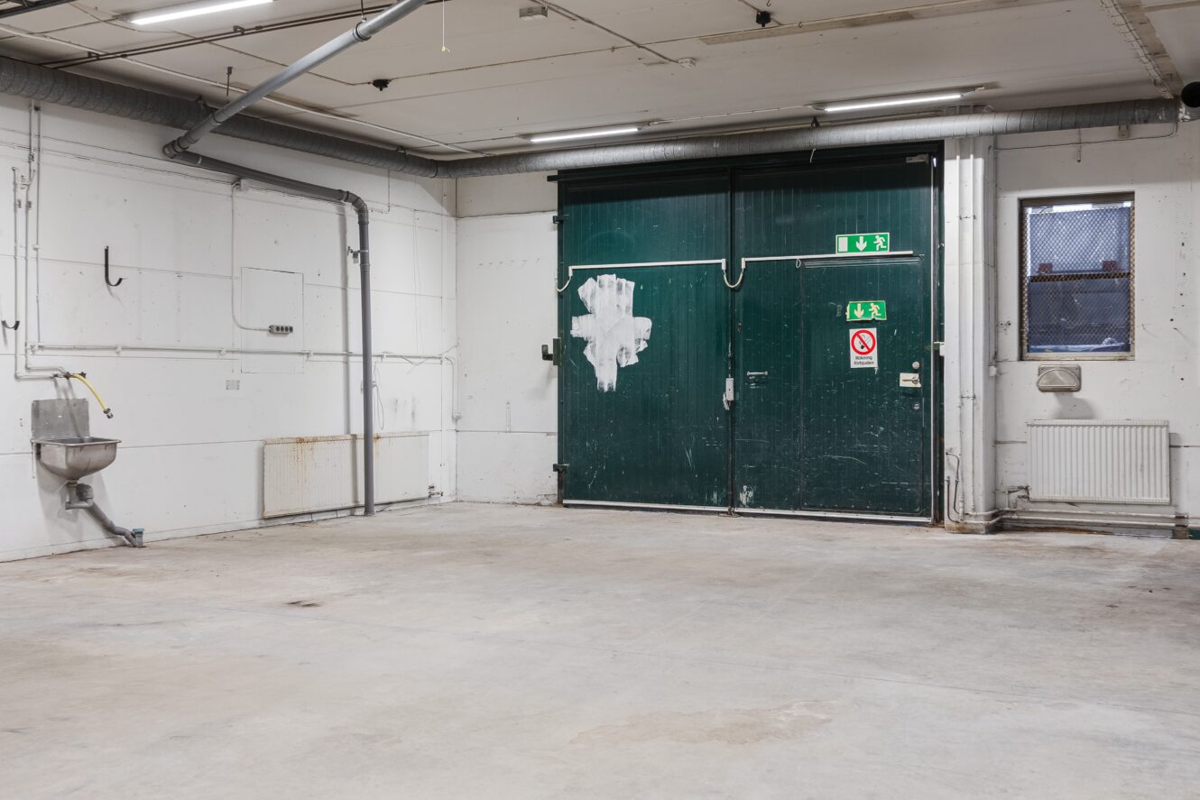 Tomt industriellt garageutrymme med betonggolv, gröna dubbeldörrar, väggmonterad diskho och synliga rör och ledningar längs vita väggar.