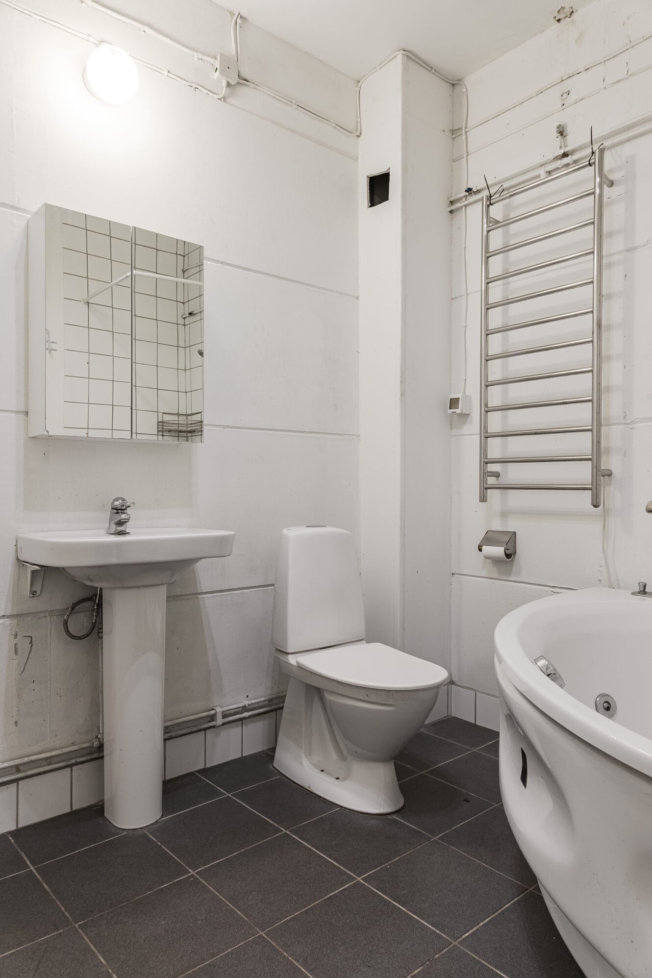 Ett modernt badrum med handfat på piedestal, väggspegel, toalett, handdukstork, toalettpappershållare och ett hörnbadkar på ett mörkt klinkergolv.