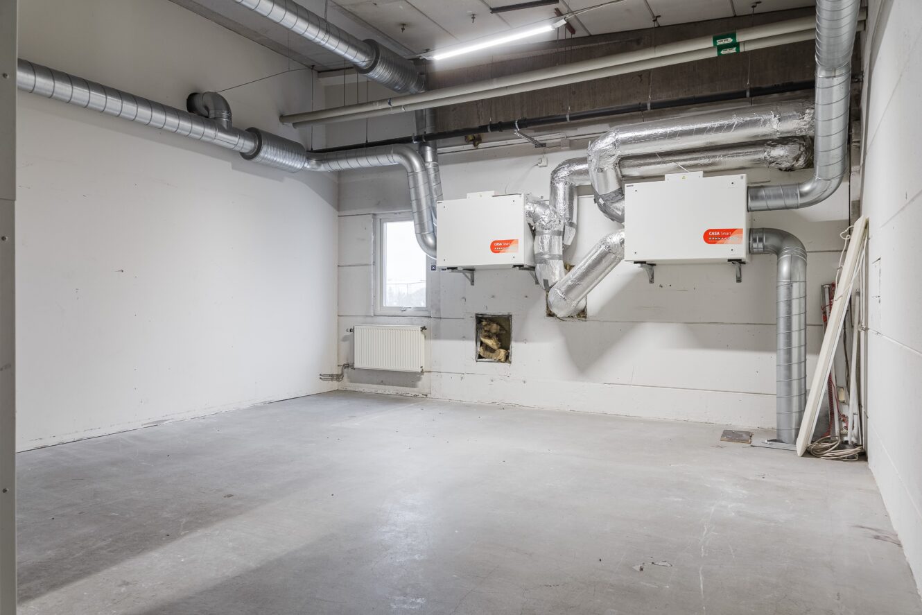 En tom industrilokal med synliga ventilationskanaler, vita väggar, en radiator under ett fönster och mekanisk utrustning monterad på väggen.