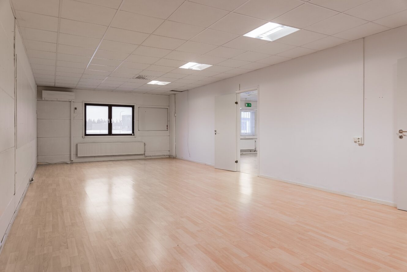 Tomt rum med ljust trägolv, vita väggar, ett fönster, taklampor och en öppen dörr som leder till ett annat liknande rum.