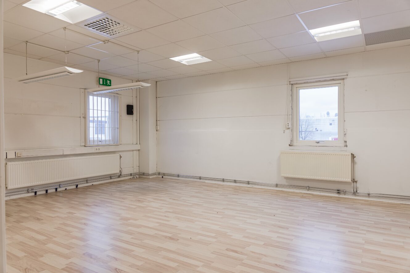 Ett tomt, väl upplyst rum med ljust trägolv, vita väggar, två fönster med galler, element, lysrör och en grön utgångsskylt ovanför dörren.