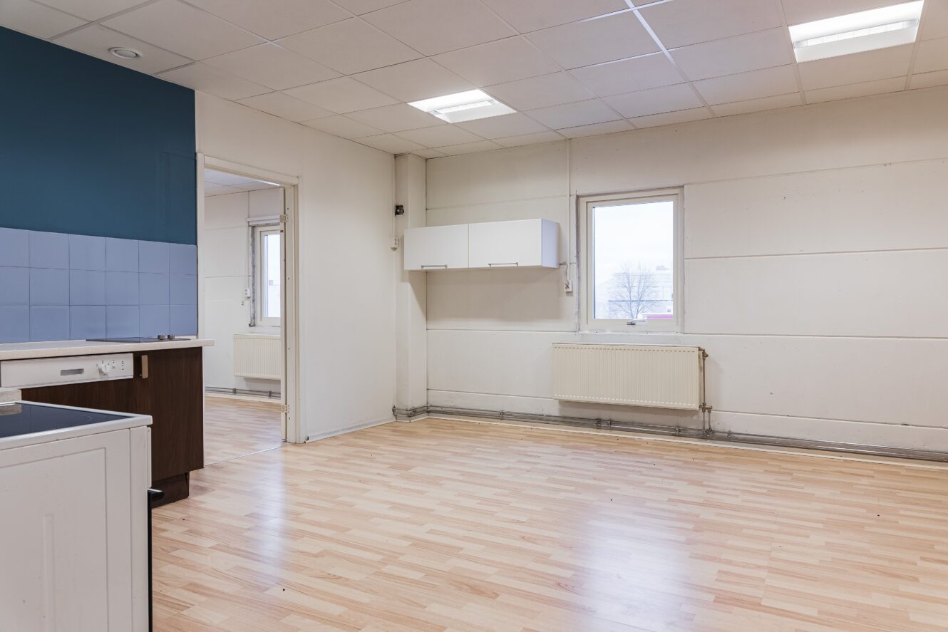 Ett tomt rum med ljust trägolv, vita väggar, en blå accentvägg, radiator, två fönster och ett litet väggskåp.