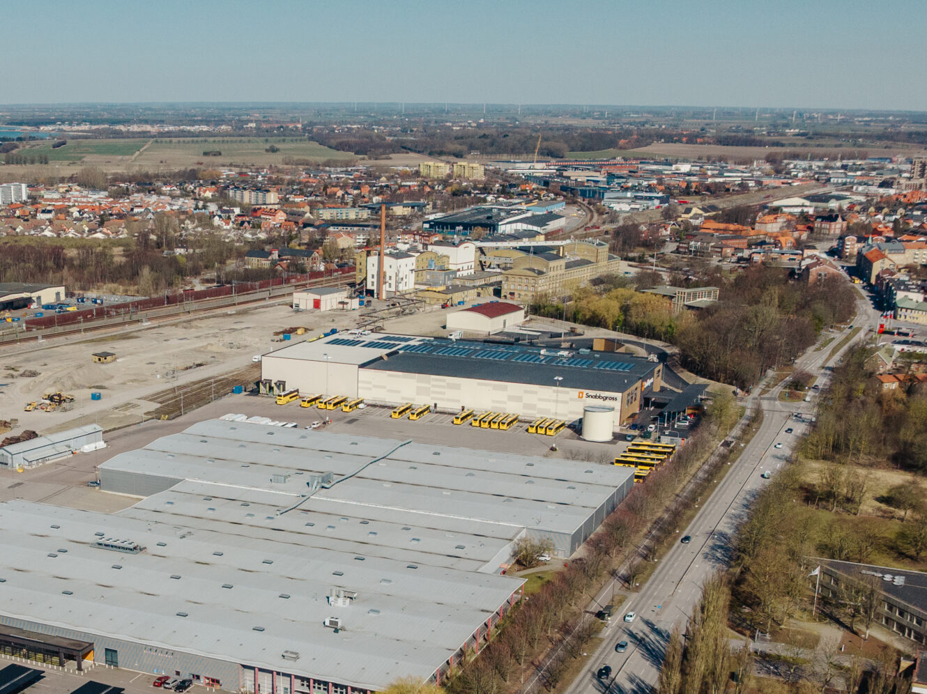 Flygfoto över ett industriområde med stora lagerbyggnader, parkeringsplatser, gula fordon och närliggande vägar, med en stad och öppna fält i bakgrunden.