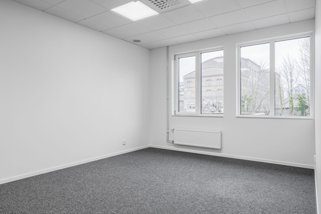 Tomt kontorsrum med vita väggar, grå heltäckningsmatta, ett stort fönster som släpper in dagsljus och ett element under fönstret.