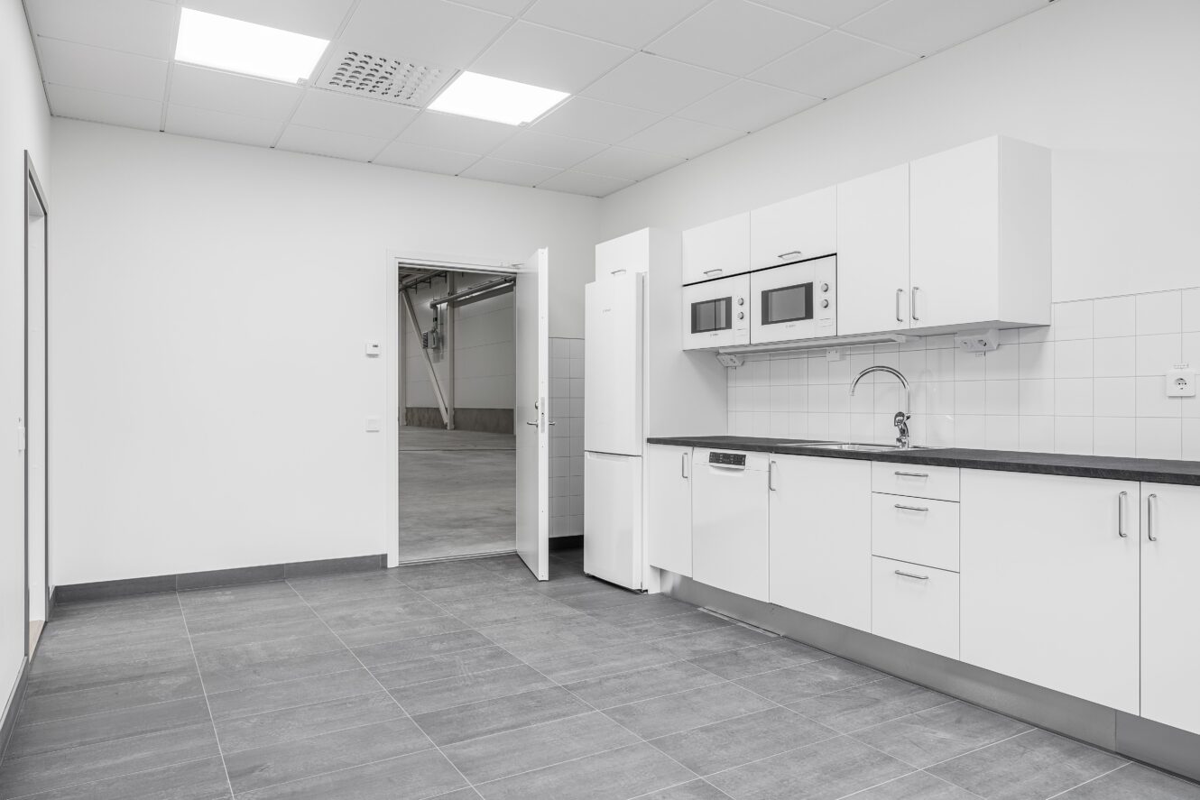 Ett modernt, tomt pentry med vita skåp, inbyggda vitvaror, grått klinkergolv och en öppen dörr som leder till ett intilliggande industriområde.