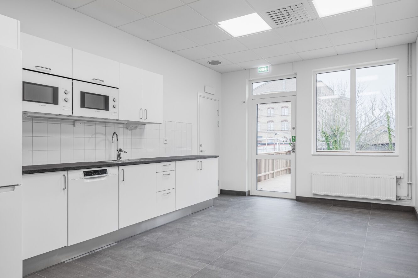 Modernt, minimalistiskt kök med vita skåp, inbyggda mikrovågsugnar, diskho och diskmaskin. Stora fönster och en glasdörr släpper in naturligt ljus. Grått klinkergolv.