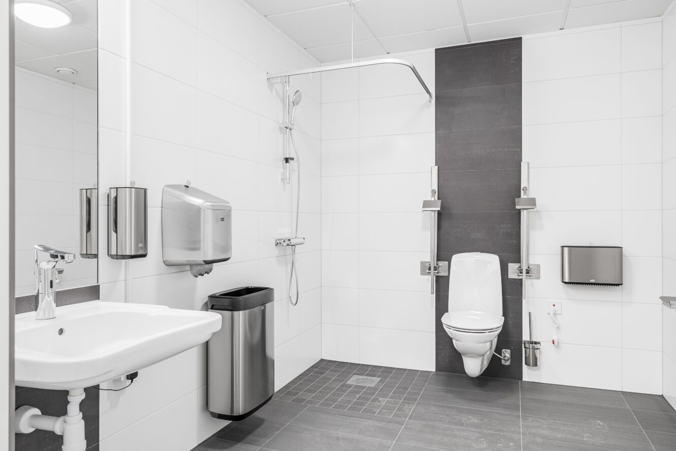 En modern, tillgänglig toalett med väggmonterad toalett, stödskenor, handfat, duschplats och sanitetsarmaturer på vita och mörkgrå kaklade väggar.