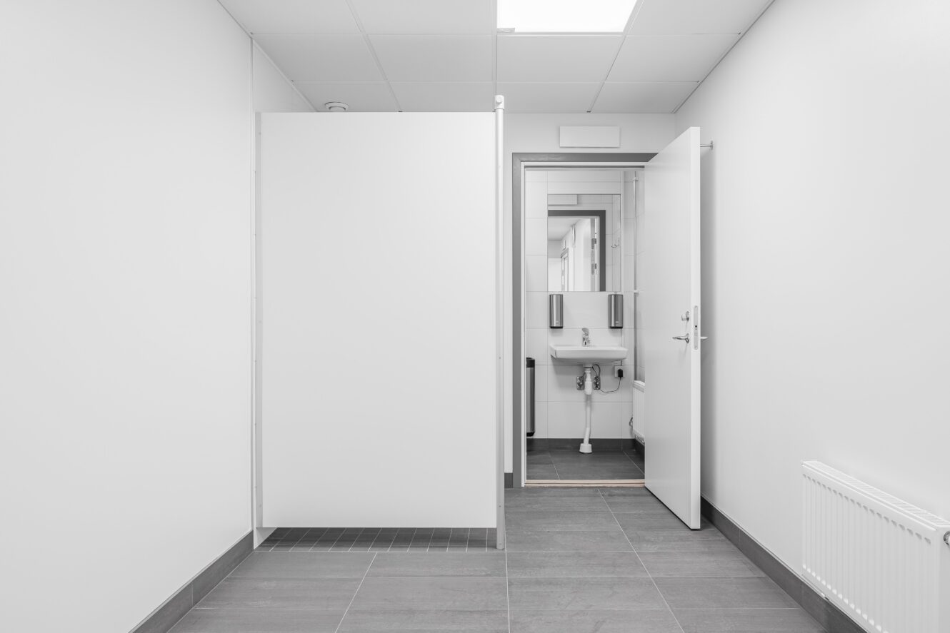 En vit toalett med öppen båsdörr, synligt handfat, spegel, tvålbehållare och en delvis öppen huvuddörr.