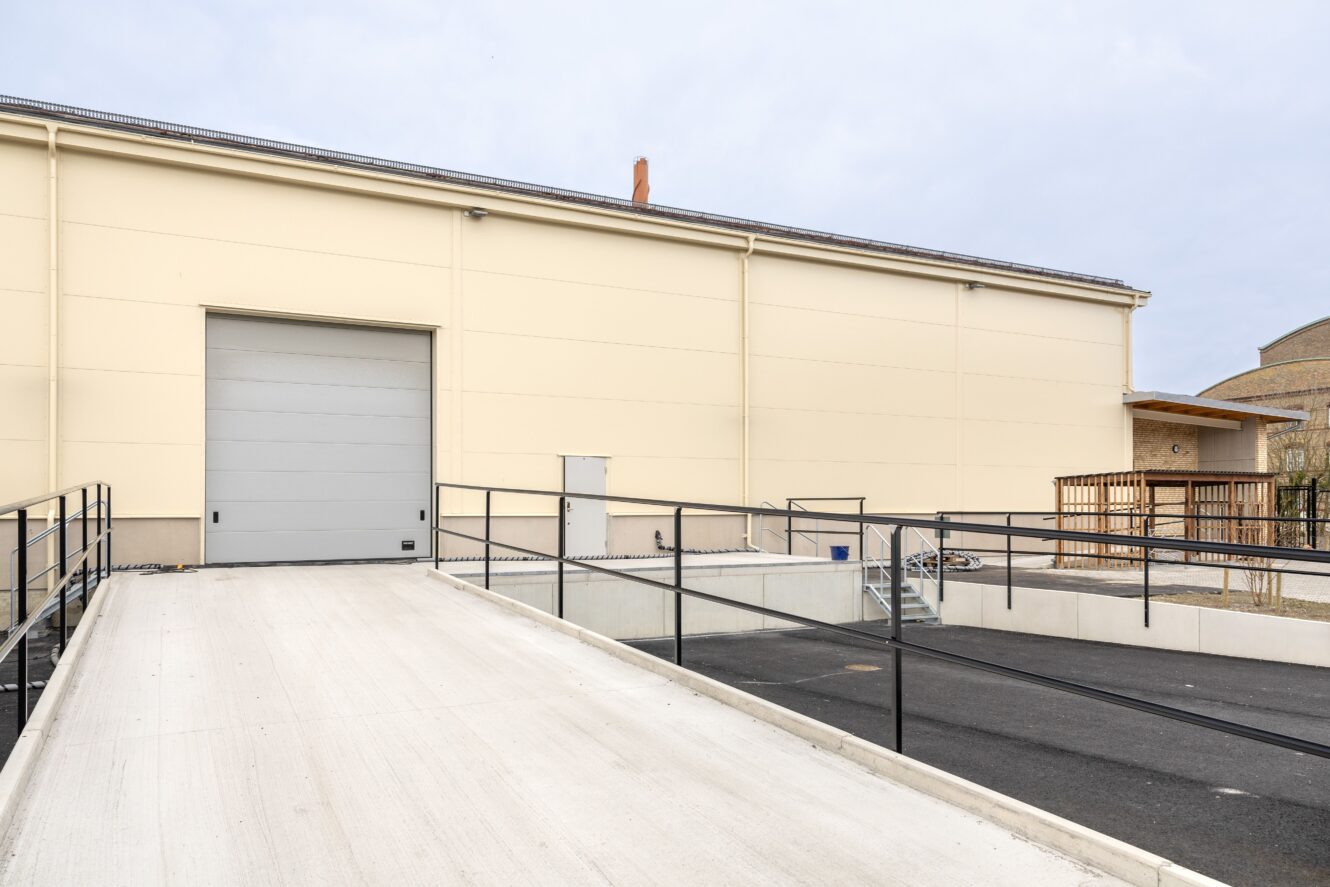 En beige lagerbyggnad med en stängd grå garageport, påfartsramp, svarta räcken och en intilliggande asfalterad yta under en molnig himmel.