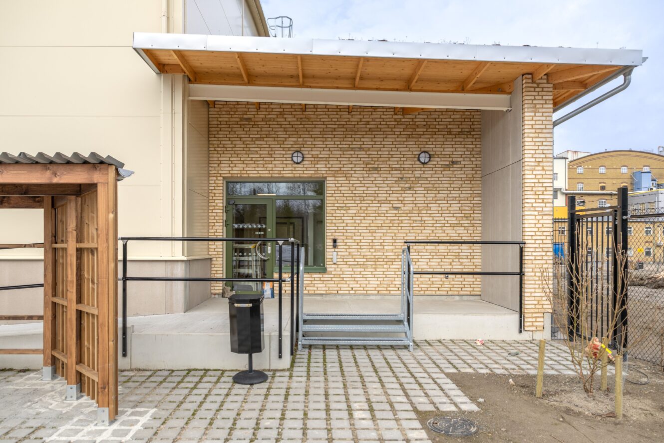 En tillgänglig entré med en metallramp, ledstänger, en tegelvägg och ett täckt tak. En soptunna och en liten träkonstruktion finns i närheten.