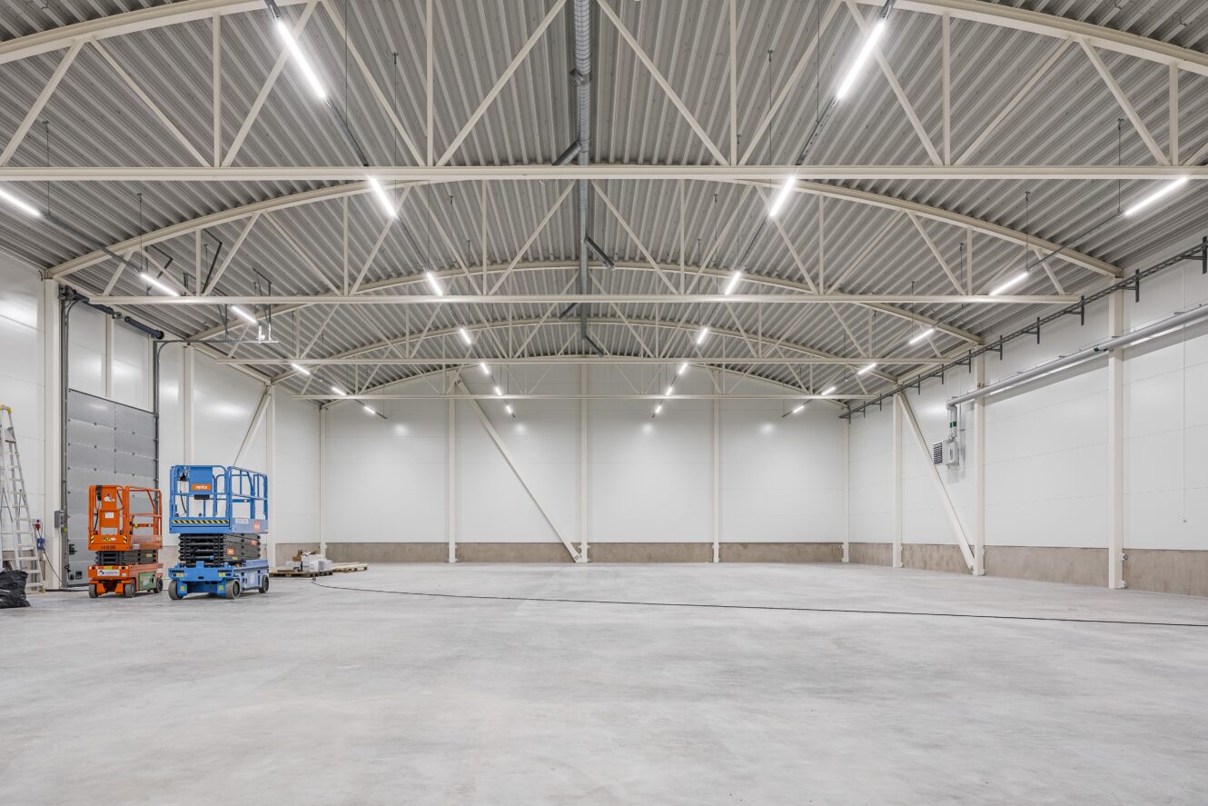 Stor, tom industrilagerlokal med högt i tak, stark takbelysning och några byggmaskiner på vänster sida.