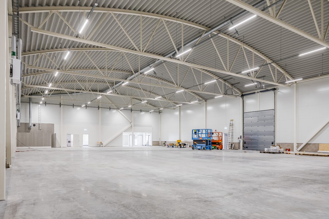 Interiör av en stor, tom industrilokal med högt i tak, stark belysning, betonggolv och byggutrustning och material längs väggarna.