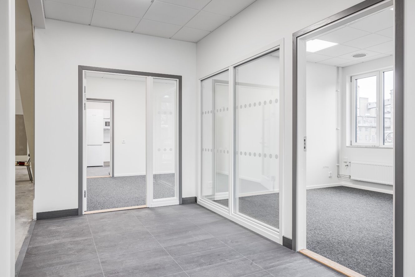 Moderna kontorslokaler med glasväggar, heltäckningsmatta i kontorsrummen, klinkergolv i korridoren, vita väggar och stora fönster med naturligt ljusinsläpp.